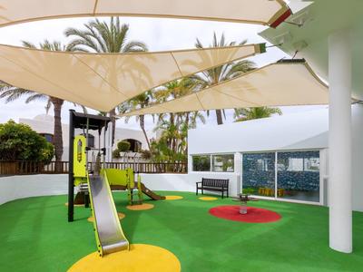 Überdachter Spielbereich mit Rutsche und Klettergerüst, umgeben von Palmen und Glaswänden.