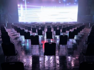Rangées de chaises dans une salle d'événement sombre avec une scène très éclairée à l'avant.