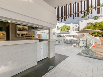 Espace extérieur moderne avec bar, sols en bois blanc, escaliers, entouré de verdure.