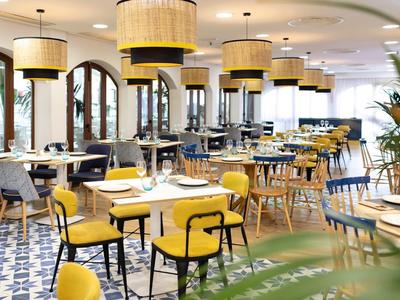 Helles Restaurant mit gelben Stühlen, runden Lampen und blau-weißem Fliesenboden.