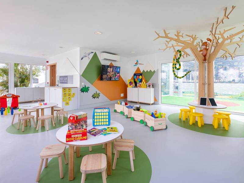 Heller, bunter Kinderspielbereich mit Tischen, Stühlen und einem dekorativen Baum im Innenraum.