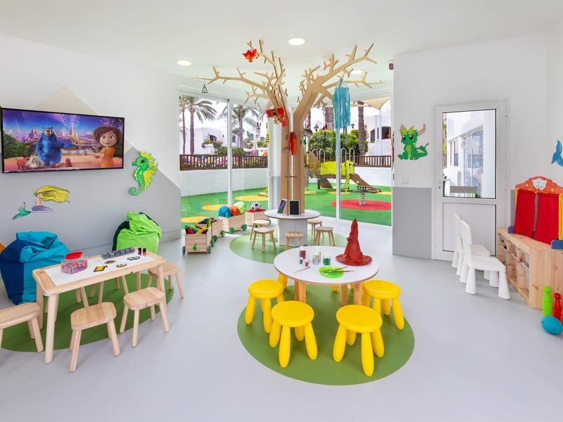 Bunter Kinderspielraum mit kleinen Tischen, Stühlen und Spielzeug in einem hellen Raum mit Fenster.