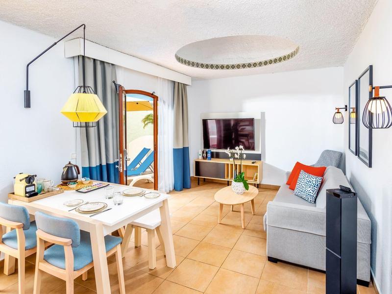 Helles Wohnzimmer mit Esstisch, Sofa, Fernseher und großem Fenster mit Vorhängen.