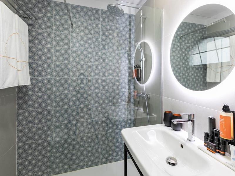 Modernes Badezimmer mit runder beleuchteter Spiegel, weißes Waschbecken und regenduschwand mit Muster