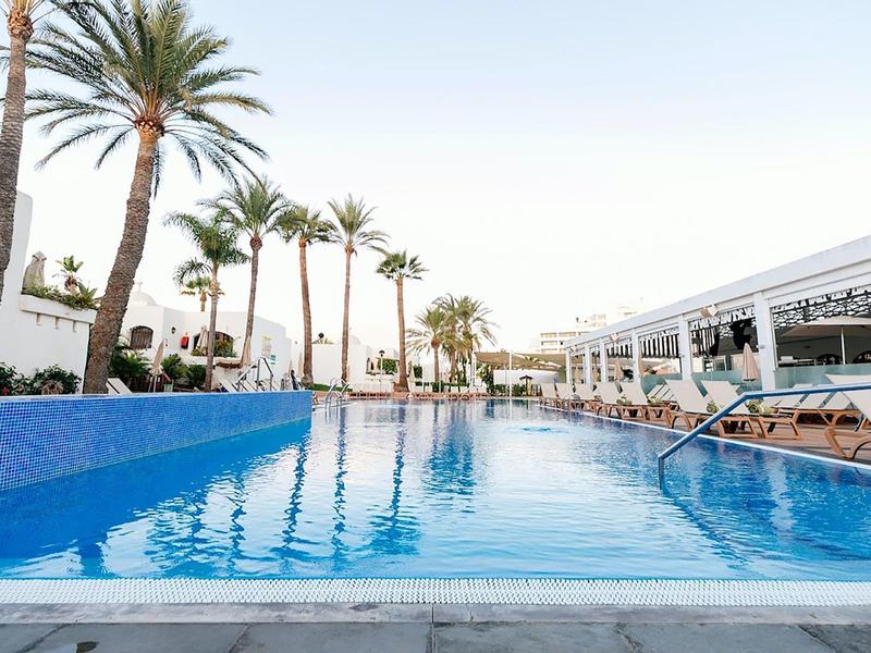 Ein klarer blauer Pool mit Palmen und Liegestühlen bei sonnigem Wetter in einer Hotelanlage.