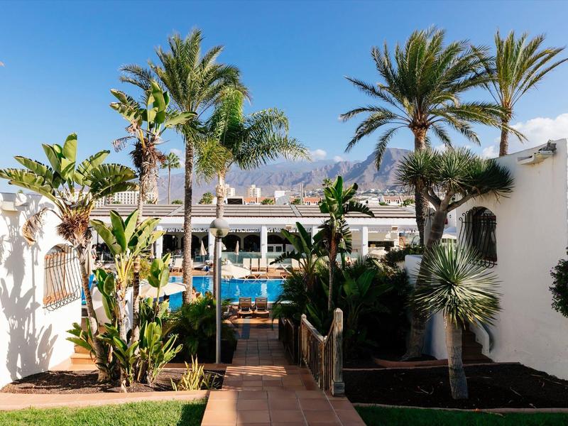 Eingang zu einem Hotel mit Palmen und einem Pool im Hintergrund bei klarem Himmel.