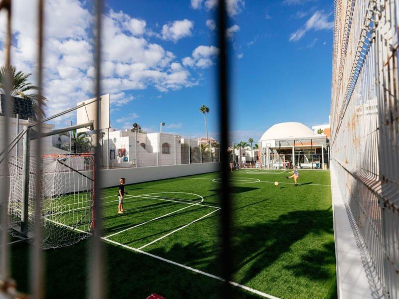 Blick durch ein Fußballtor auf einen grünen Spielfeldrasen neben einem weißen Hotelgebäude unter blauem Himmel.