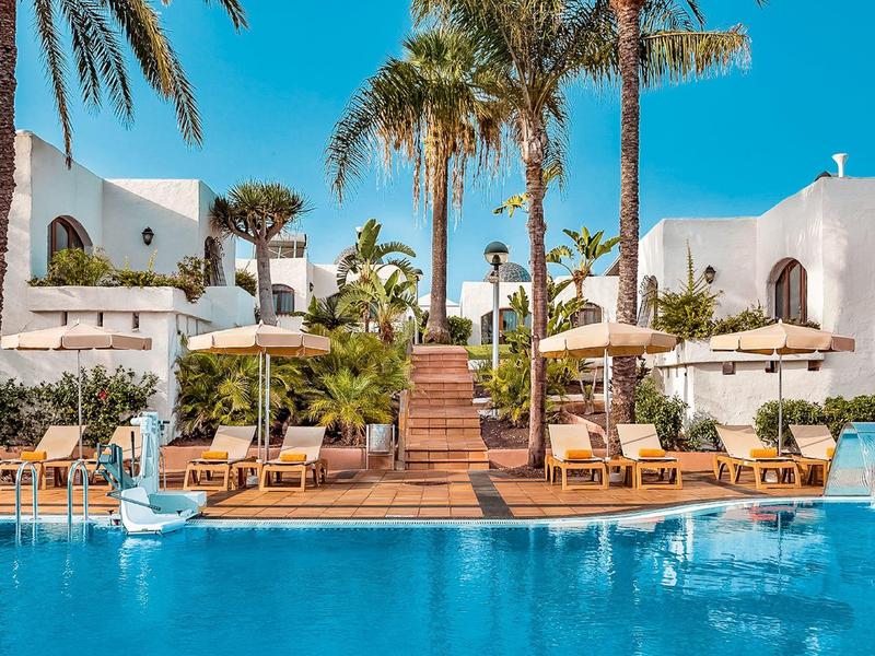 Inselresort mit blauem Pool, weißen Gebäuden, Palmen und Sonnenschirmen unter klarem Himmel.