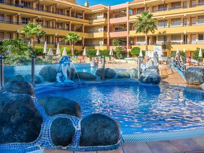 Piscina del hotel con rocas, juegos de agua y sombrillas en un complejo hotelero amarillo.