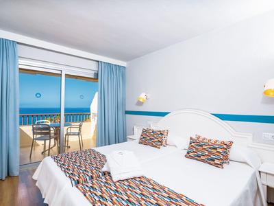 Habitación de hotel luminosa con cama doble, cojines coloridos y terraza con vista al mar.