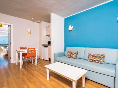 Área de estar luminosa con sofá azul, mesa blanca y pequeña mesa de comedor con dos sillas naranjas.