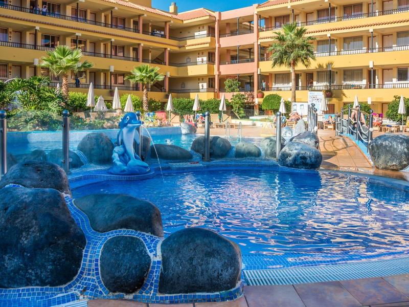 Piscina del hotel con rocas, juegos de agua y sombrillas en un complejo hotelero amarillo.