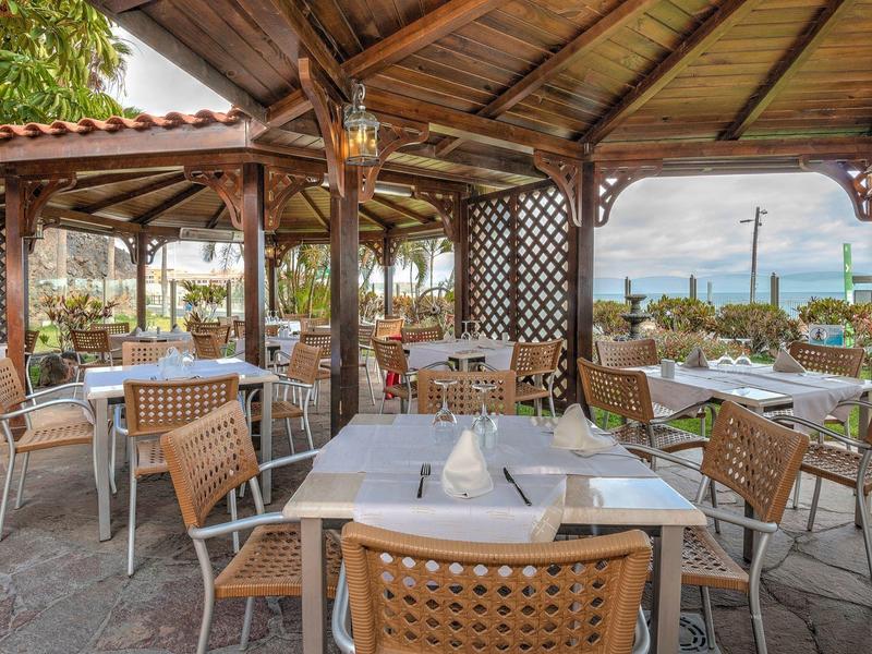 Terraza de restaurante al aire libre con mesas y sillas, decorada con luces y plantas bajo techo de madera.