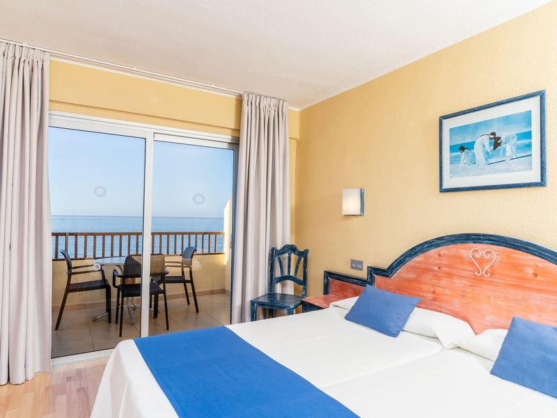 Habitación de hotel luminosa con cama doble, ropa de cama azul y blanca y balcón con vista al mar.