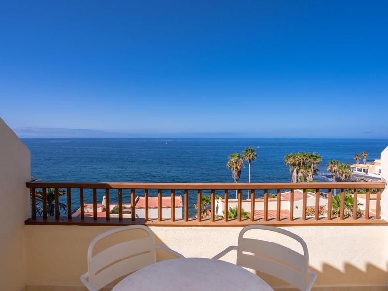 Balcón con mesa y sillas con vista al mar azul y palmeras bajo cielo despejado.