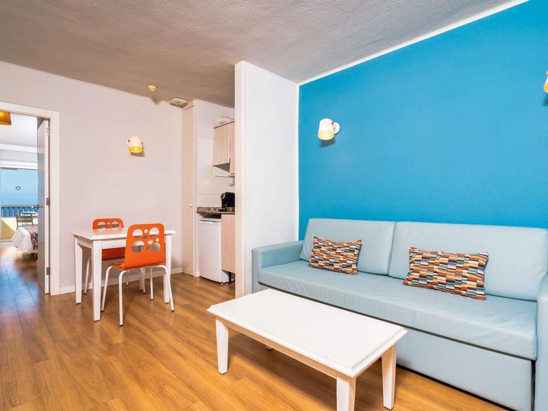 Área de estar luminosa con sofá azul, mesa blanca y pequeña mesa de comedor con dos sillas naranjas.