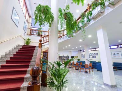 Světlá hotelová hala s mramorovou podlahou, zelenými rostlinami a schodištěm s červeným kobercem.