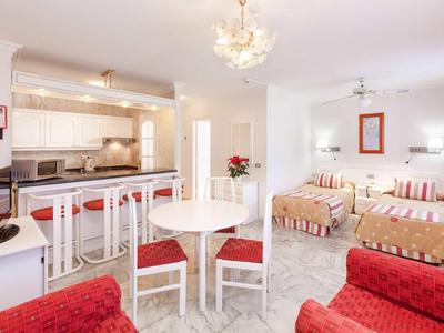 Hell eingerichtetes Zimmer mit kleiner Küche, Essbereich, zwei Betten und roten Sitzmöbeln.