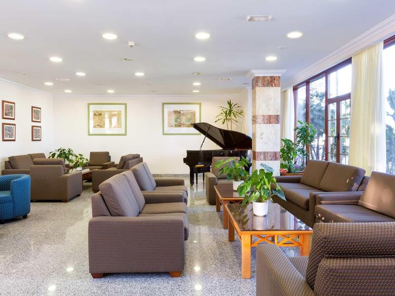 Heller Wartesaal mit braunen Sesseln, Holztischen, großem Fenster und schwarzem Flügelklavier.