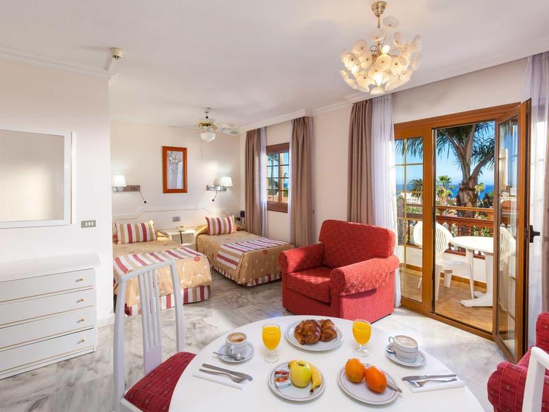 Helles Wohnzimmer mit Esstisch, Sesseln, Doppelbett und Balkon mit Meerblick und Palmen.