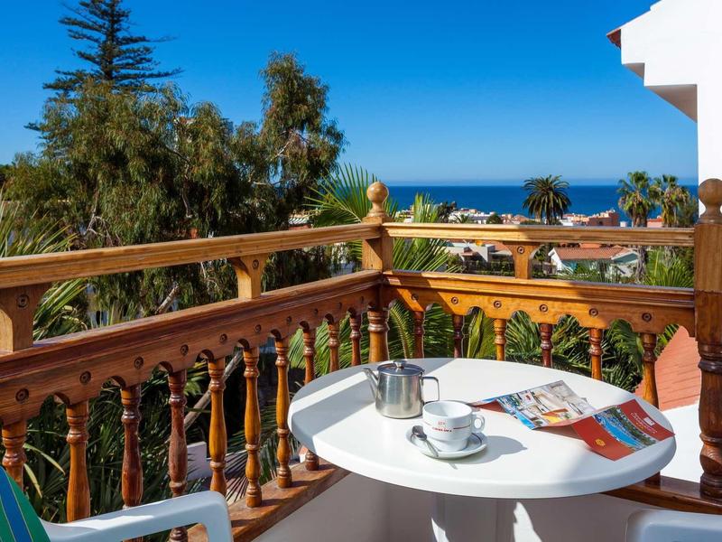 Balkon mit weißem Tisch, Tee, Zeitschrift, Holzgeländer und Blick auf Meer und Palmen.