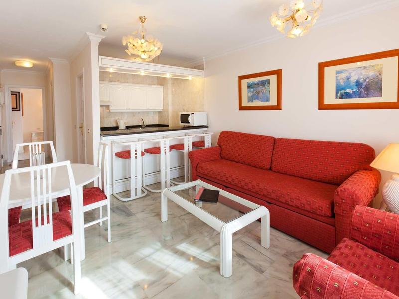 Helles Wohnzimmer mit rotem Sofa, Glas-Couchtisch, weißer Essgruppe und moderner Kitchenette im Hintergrund.