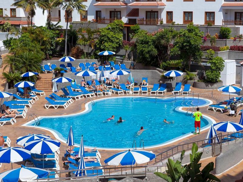 Runder Pool mit blauen Liegestühlen und Sonnenschirmen, umgeben von weißen Gebäuden und Palmen.