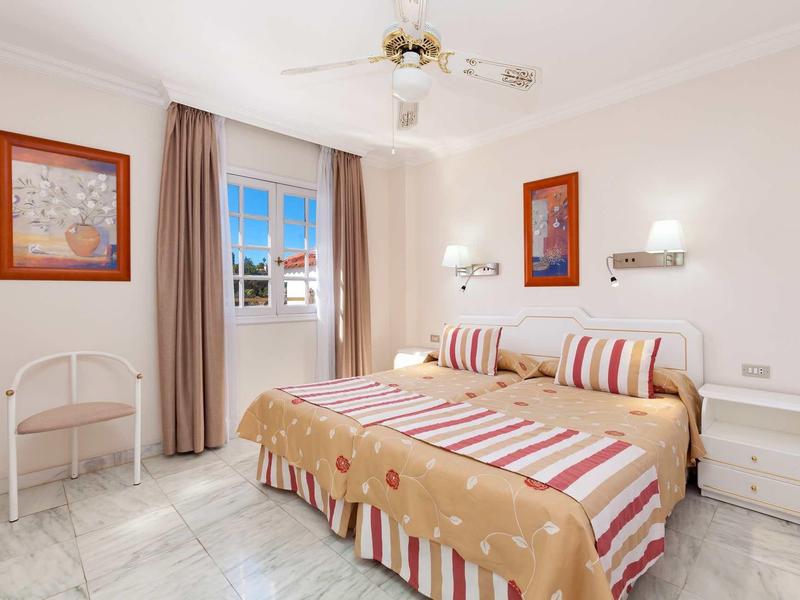 Helles Schlafzimmer mit großem Bett, zweifarbigem Bettbezug, zwei Nachttischen und Fenster mit Vorhang.