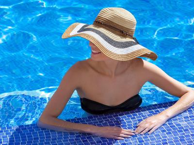 Frau mit großem Sonnenhut und schwarzem Bikini lehnt entspannt am Beckenrand in blauem Wasser.