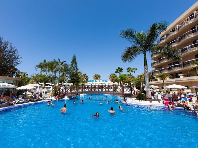 Grande piscine d'hôtel avec des personnes, des palmiers et un ciel bleu clair par temps ensoleillé.