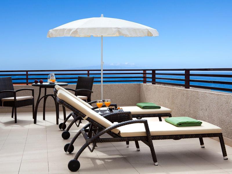 Dwa leżaki z zielonymi poduszkami pod białym parasolem na tarasie z widokiem na morze.