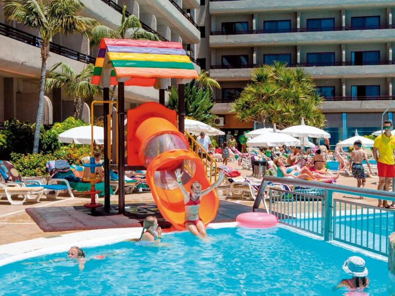 Dzieci bawią się na zjeżdżalni w hotelowym basenie z leżakami i parasolami.