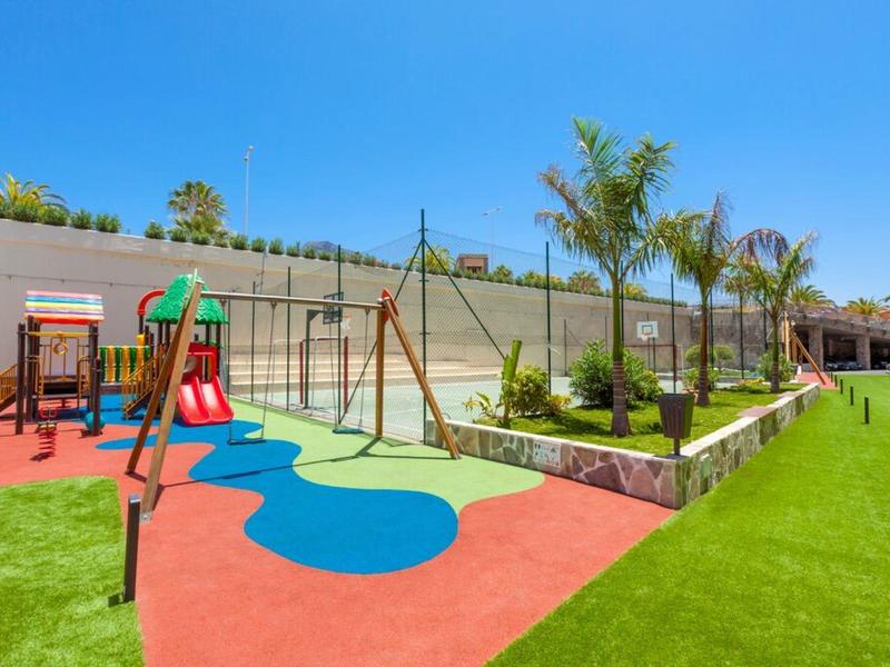 Plac zabaw dla dzieci z zjeżdżalniami, huśtawkami i kolorową gumową matą obok trawiastego terenu.