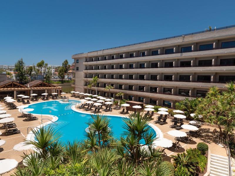 Hotel z dużym basenem, leżakami i parasolami, otoczony roślinnością.