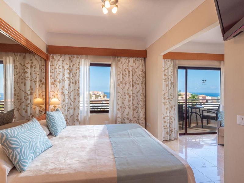 Jasny pokój hotelowy z podwójnym łóżkiem, balkonem i widokiem na morze w słoneczny dzień.