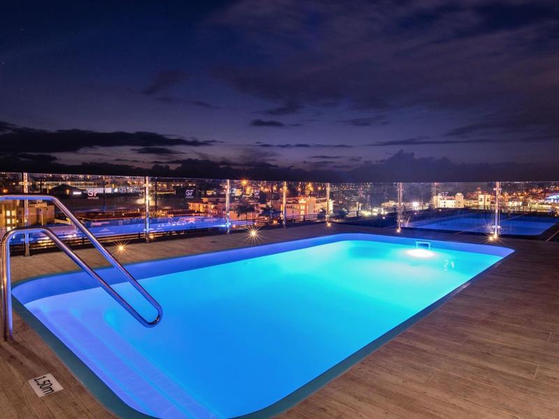 Oświetlony basen na dachu nocą z widokiem na miasto w tle.