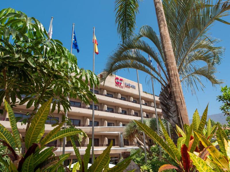 Budynek hotelowy z wieloma balkonami otoczony palmami i roślinami pod czystym niebem.