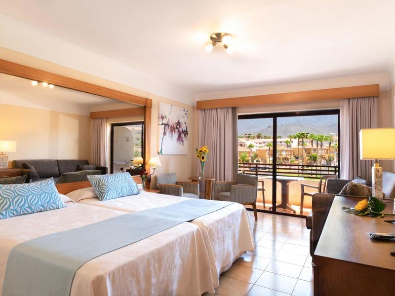 Jasny pokój hotelowy z podwójnymi łóżkami, jasnymi zasłonami i balkonem z widokiem.