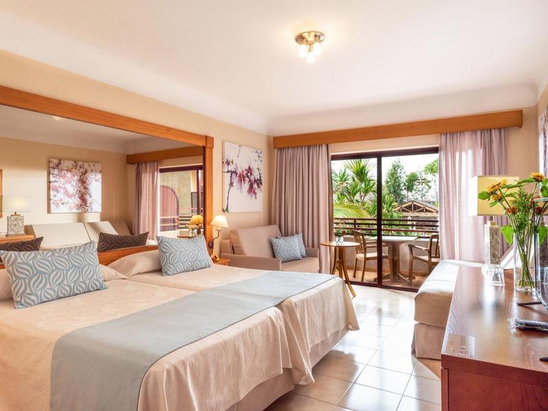 Jasny pokój hotelowy z dwoma łóżkami pojedynczymi, częścią wypoczynkową i balkonem z widokiem na ogród.