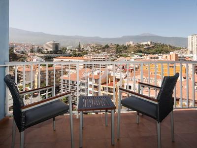 Balkon mit zwei Stühlen und Tisch, rote Fliesen, Blick auf Stadt und Berge im Hintergrund.