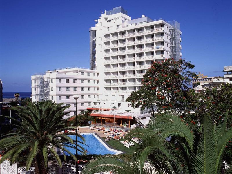 Haut bâtiment d'hôtel blanc avec piscine et palmiers sous un ciel bleu clair.