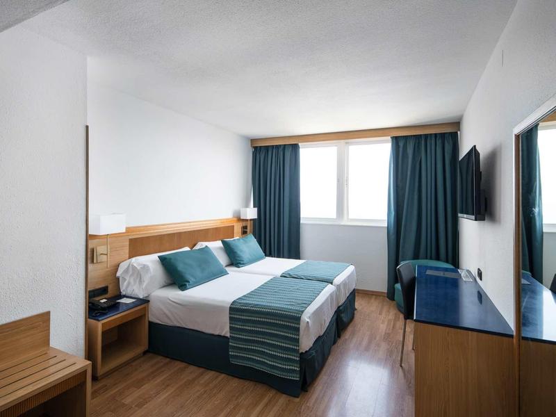 Chambre d'hôtel moderne avec lit double, sol en bois et grande fenêtre avec rideaux bleus.