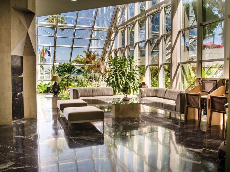 Salon d'hôtel lumineux avec mur en verre, plantes et sièges modernes sur sol en marbre.
