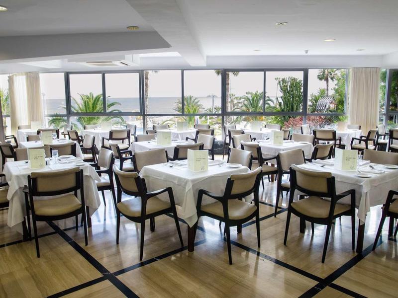 Salle à manger lumineuse avec nappes blanches, nombreuses chaises et grandes fenêtres en arrière-plan.