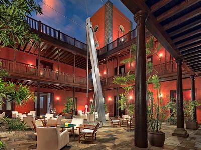 Innenhof mit roten Wänden, Holzbalkonen, Pflanzen und bequemen weißen Sitzmöbeln bei Abendlicht.