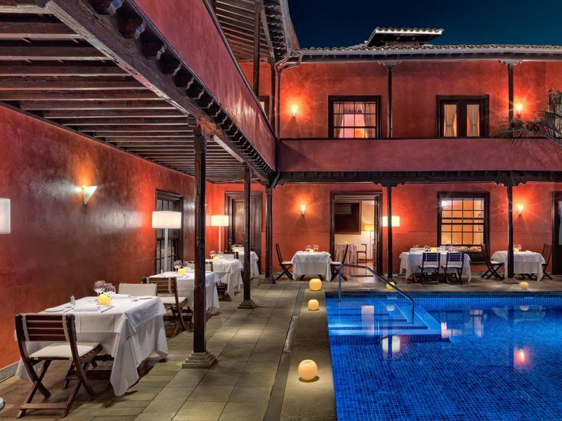 Hotelowy taras z nakrytymi stolikami i oświetlonym basenem w nocy.