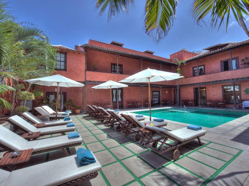 Strefa basenowa z leżakami i parasolami przed czerwonym budynkiem hotelu w słoneczny dzień.