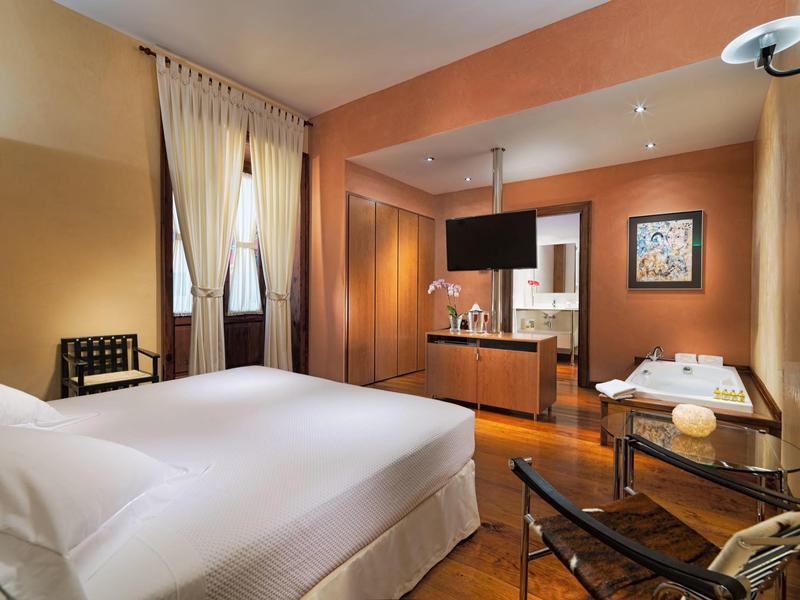 Nowoczesny pokój hotelowy z podwójnym łóżkiem, aneksem kuchennym i stołem w ciepłych kolorach.