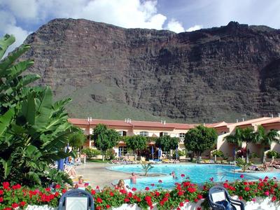 Hotel mit blauem Pool, roten Blumen und hohen Felsen im Hintergrund unter blauem Himmel.