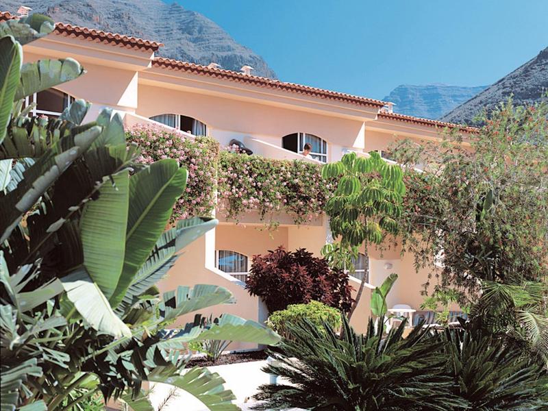 Un bâtiment hôtelier à l'architecture méditerranéenne, entouré de verdure luxuriante et de montagnes.